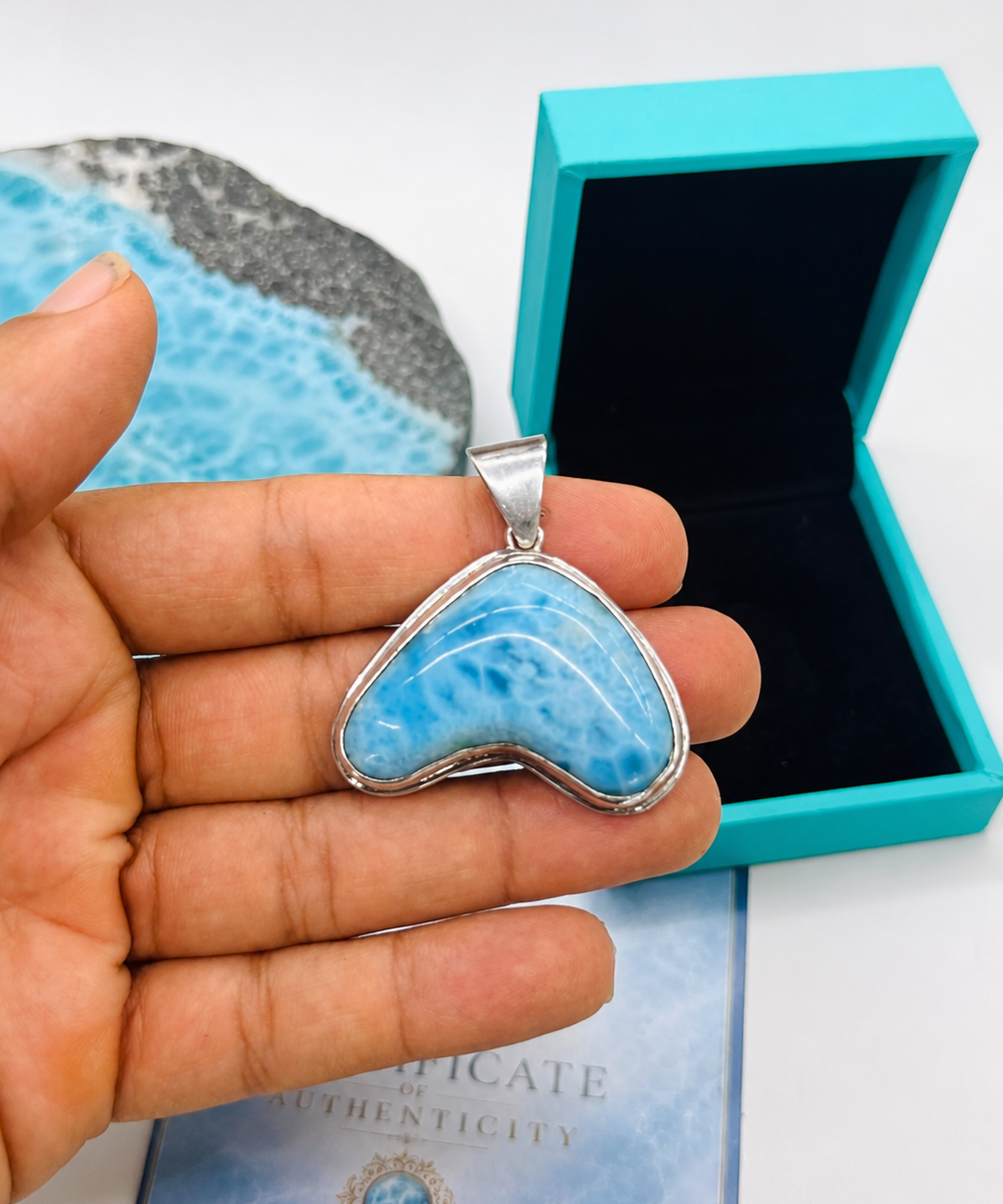 Heart-shaped blue stone pendant held by a hand with turquoise box and card in the background