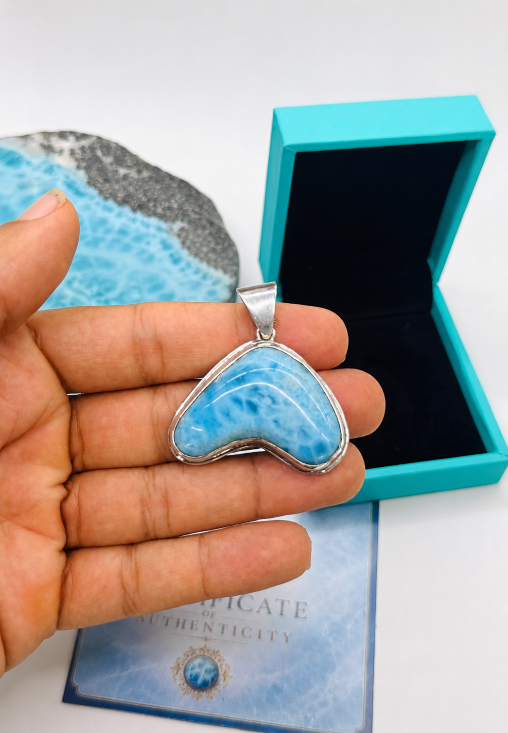 Heart-shaped blue stone pendant held by a hand with turquoise box and card in the background