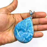A teardrop-shaped Larimar pendant with a silver bezel setting, held in the palm of a hand against a plain white background.