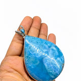 A hand holding a teardrop-shaped blue marbled stone pendant with a silver-toned bezel and bail, shown against a plain white background.
