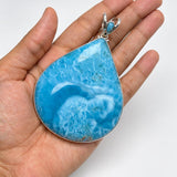A hand holding a teardrop-shaped Larimar pendant with a silver bezel and bail, set against a plain white background.