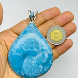 A large teardrop-shaped turquoise pendant with a silver bail, held in a person's hand next to a coin for scale, against a plain light gray background.