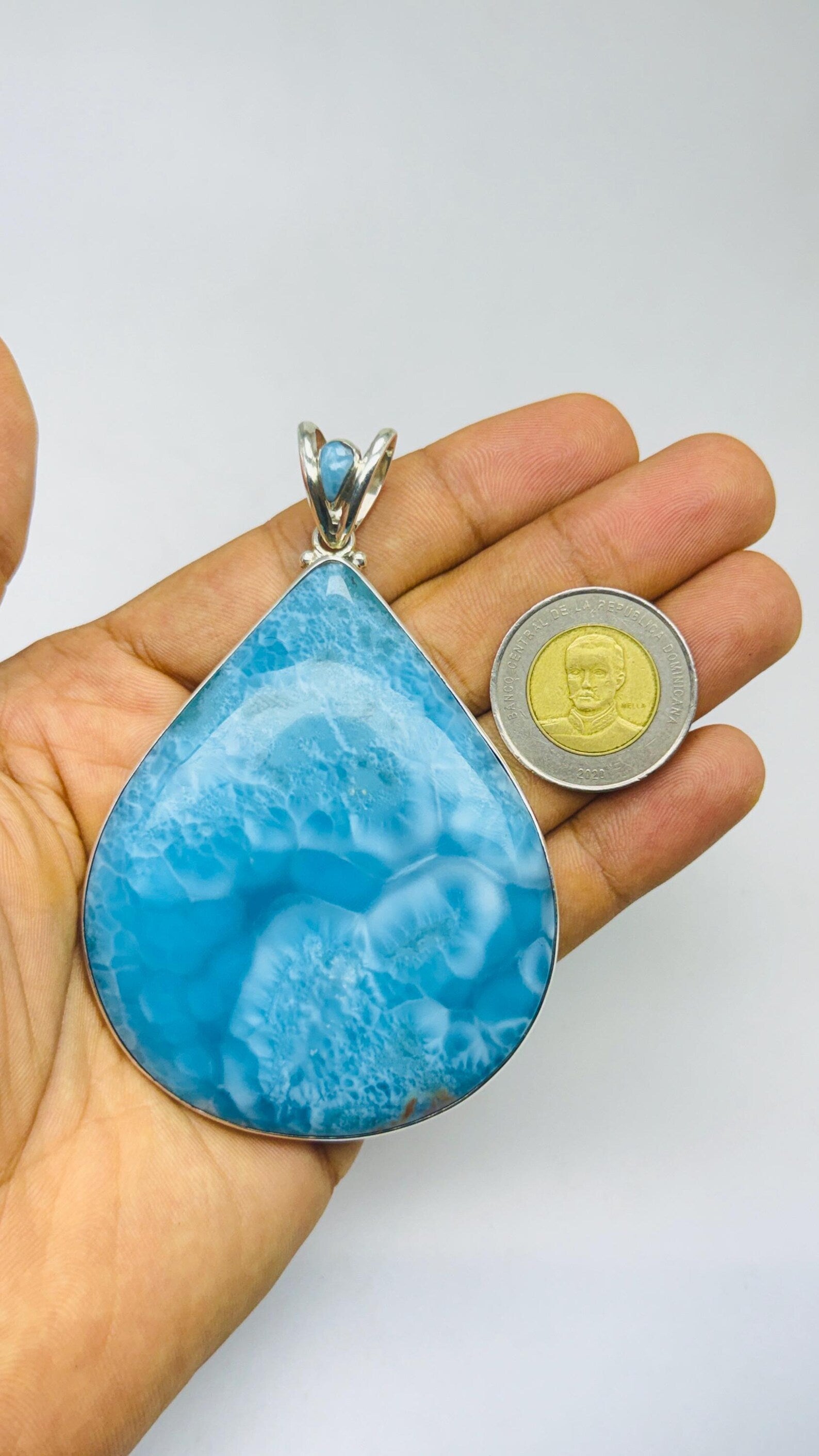 A large teardrop-shaped turquoise pendant with a silver bail, held in a person's hand next to a coin for scale, against a plain light gray background.