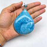 A hand holding a large teardrop-shaped turquoise pendant with a silver-toned setting against a plain white background.