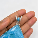 A close-up of a hand holding a large teardrop-shaped Larimar pendant with a silver bail and a small turquoise accent.