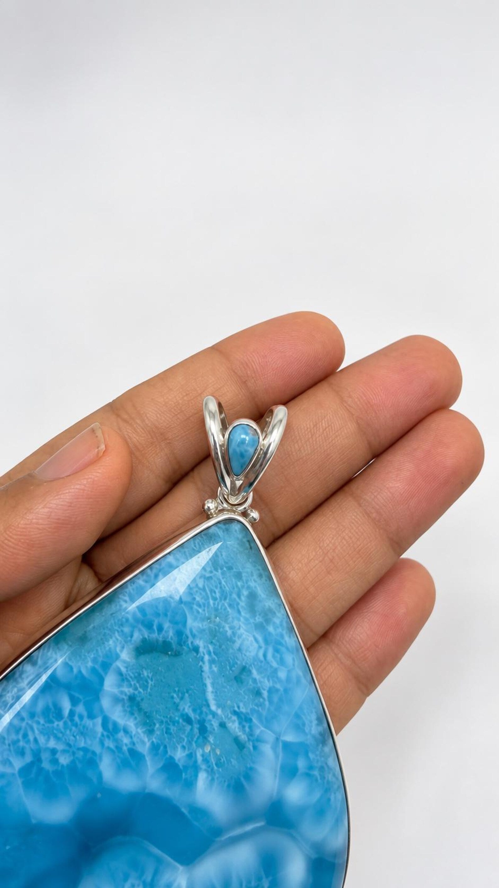 A close-up of a hand holding a large teardrop-shaped Larimar pendant with a silver bail and a small turquoise accent.