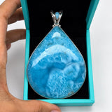 A teardrop-shaped blue marbled gemstone pendant in a silver setting, presented inside an open turquoise jewelry box held by a hand against a white background.