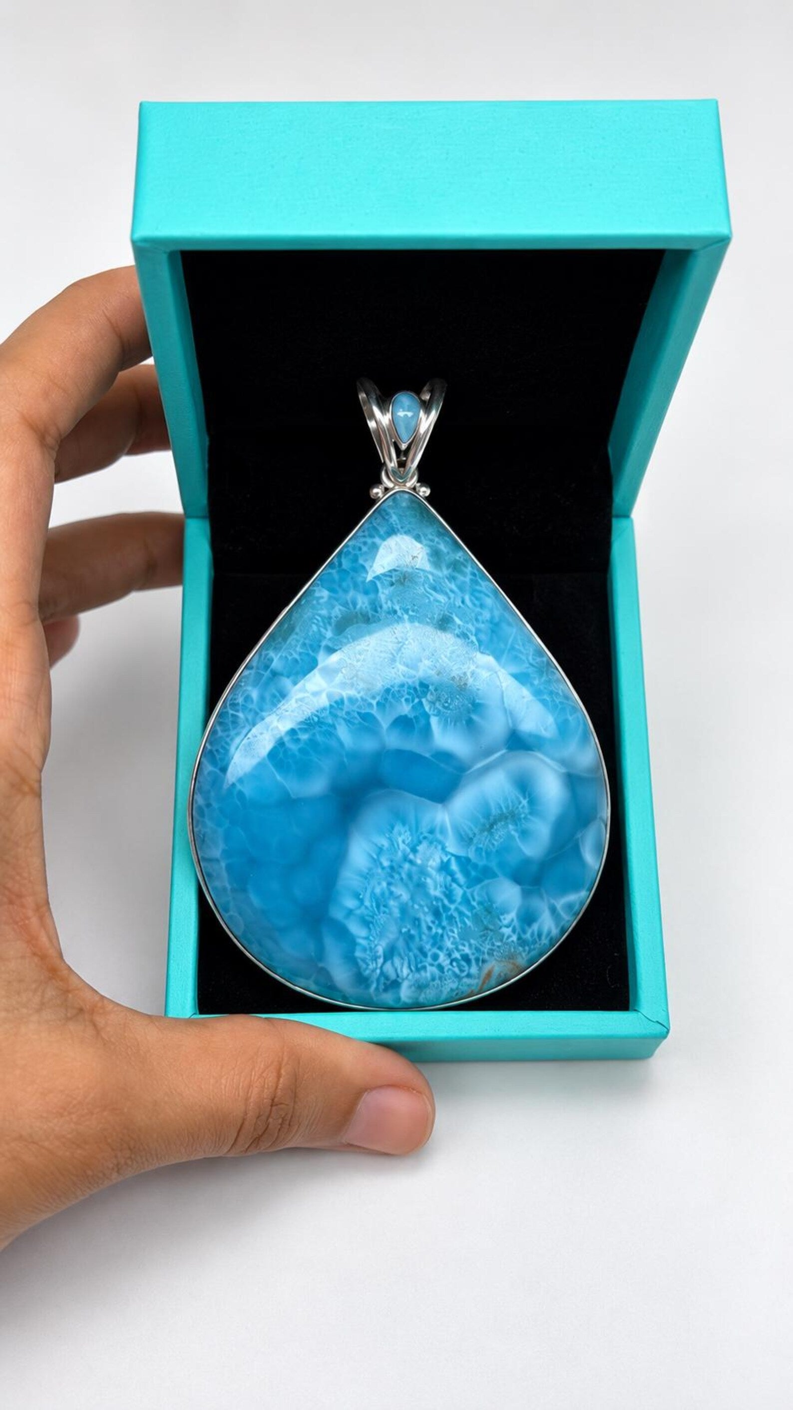 A teardrop-shaped blue marbled gemstone pendant in a silver setting, presented inside an open turquoise jewelry box held by a hand against a white background.