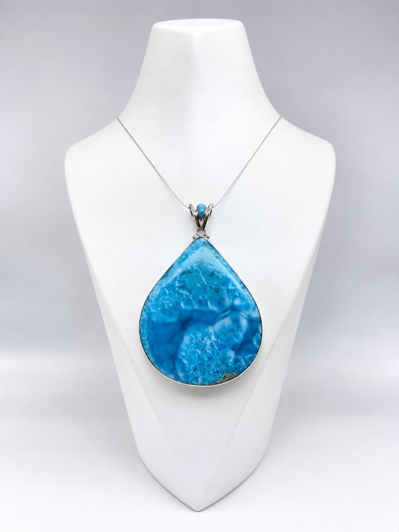 A teardrop-shaped turquoise pendant necklace displayed on a white jewelry bust. The pendant features natural marbled blue and white patterns, set in a silver-toned bezel with a delicate silver chain.