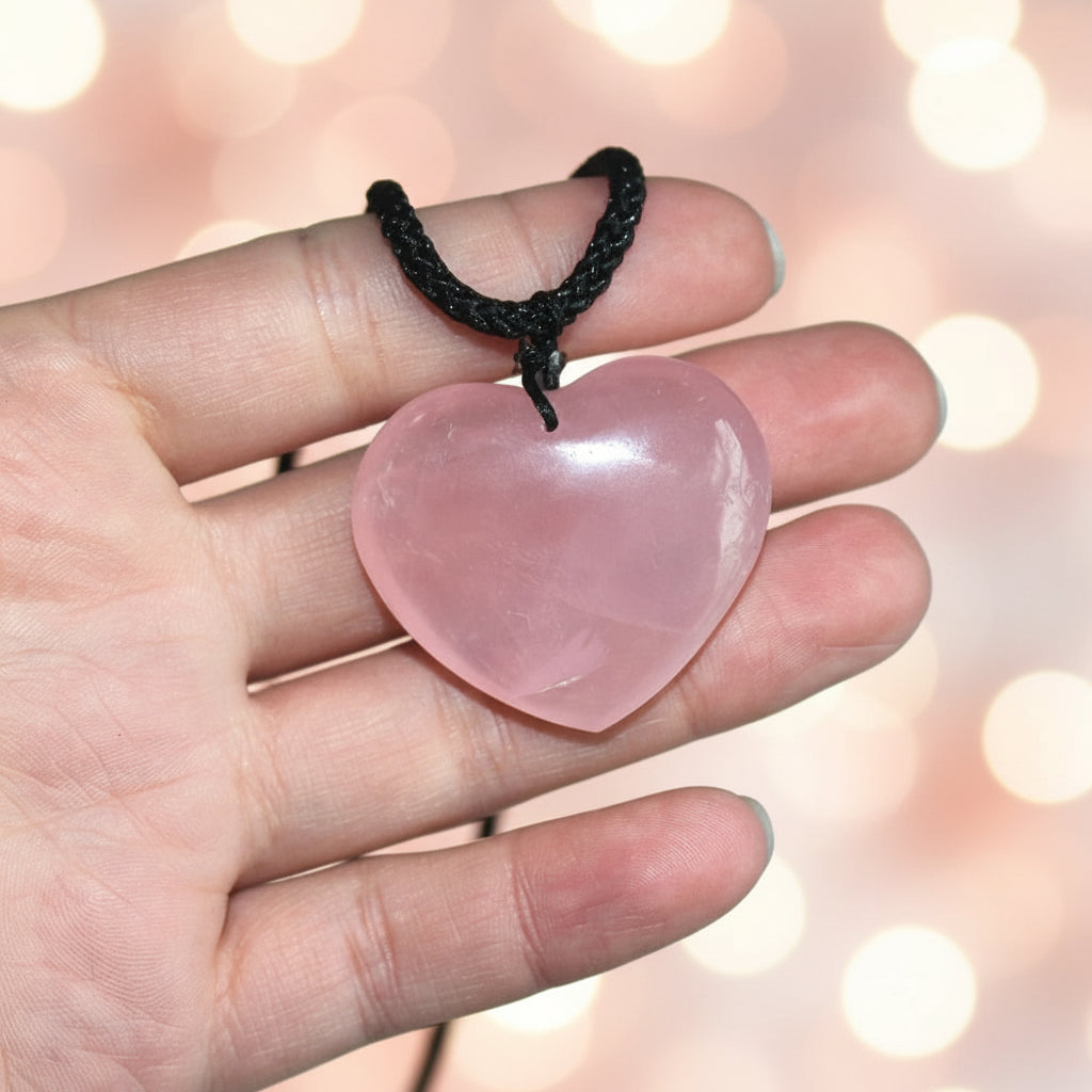 Heart-shaped pink stone pendant on a black cord held in a hand against a neutral background
