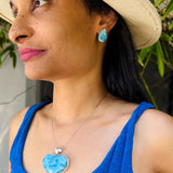 A woman wearing a light beige straw hat, a blue ribbed tank top, and a silver Larimar heart pendant necklace with a light blue marbled stone. She is also wearing a matching light blue teardrop earring.