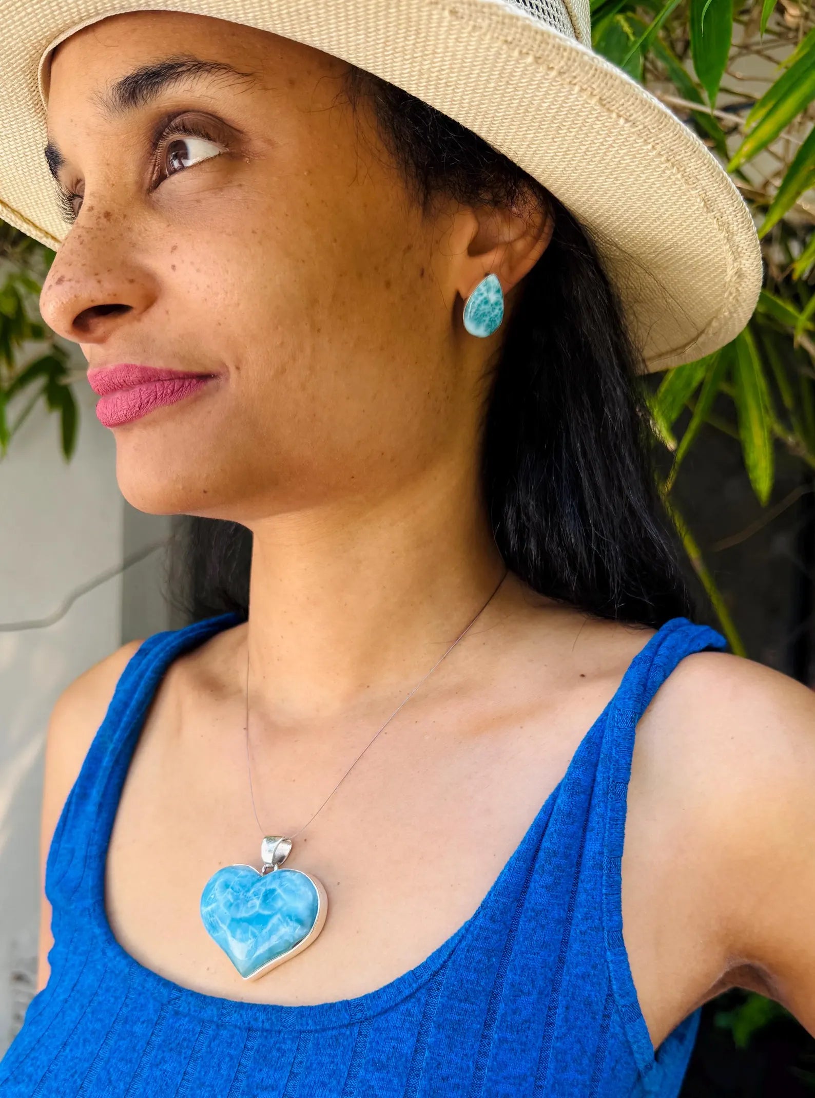 A woman wearing a light beige straw hat, a blue ribbed tank top, and a silver Larimar heart pendant necklace with a light blue marbled stone. She is also wearing a matching light blue teardrop earring.