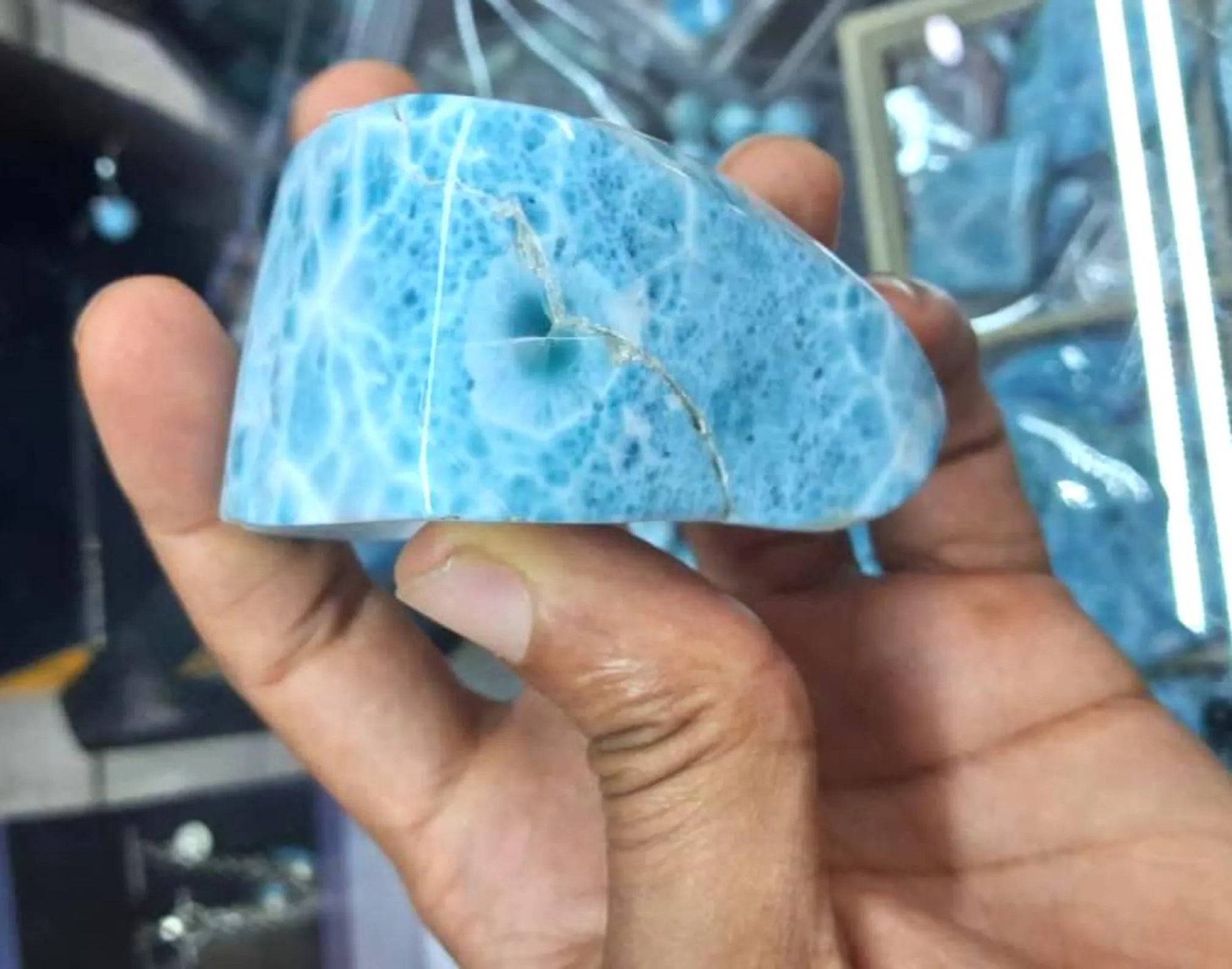 A hand holding a polished blue stone or crystal with natural marbled patterns and a visible crack, photographed against a blurred indoor background.