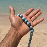 A person's hand holding a Larimar Bead Necklace beads on a sandy beach with the ocean in the background.