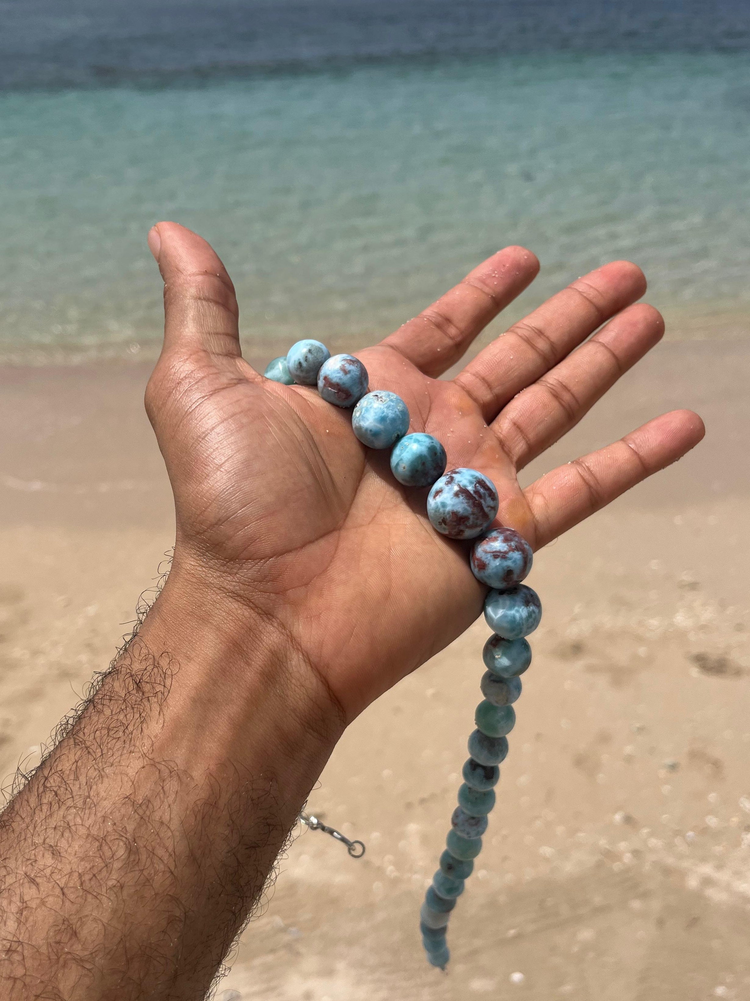 A person's hand holding a Larimar Bead Necklace beads on a sandy beach with the ocean in the background.