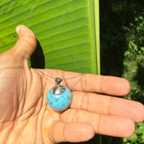 A person's hand holding a small blue stone pendant necklace with a silver bail, set against a background of a large green banana leaf outdoors.