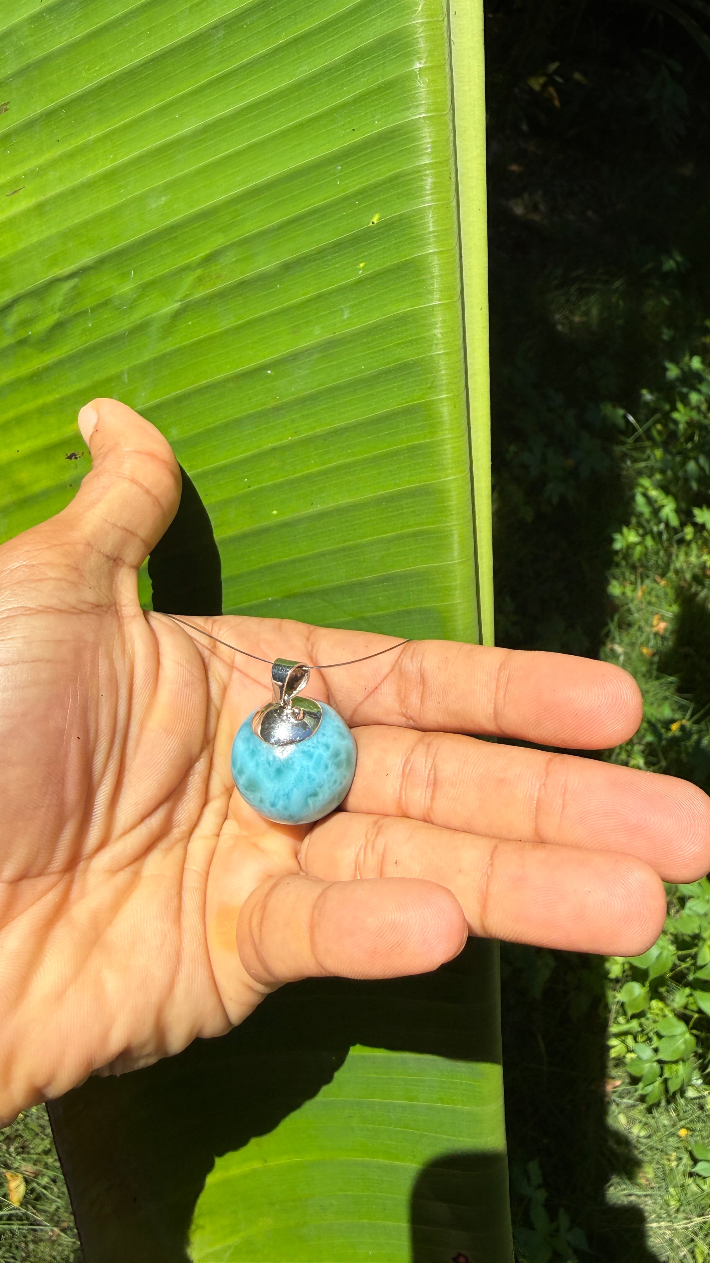 A person's hand holding a small blue stone pendant necklace with a silver bail, set against a background of a large green banana leaf outdoors.