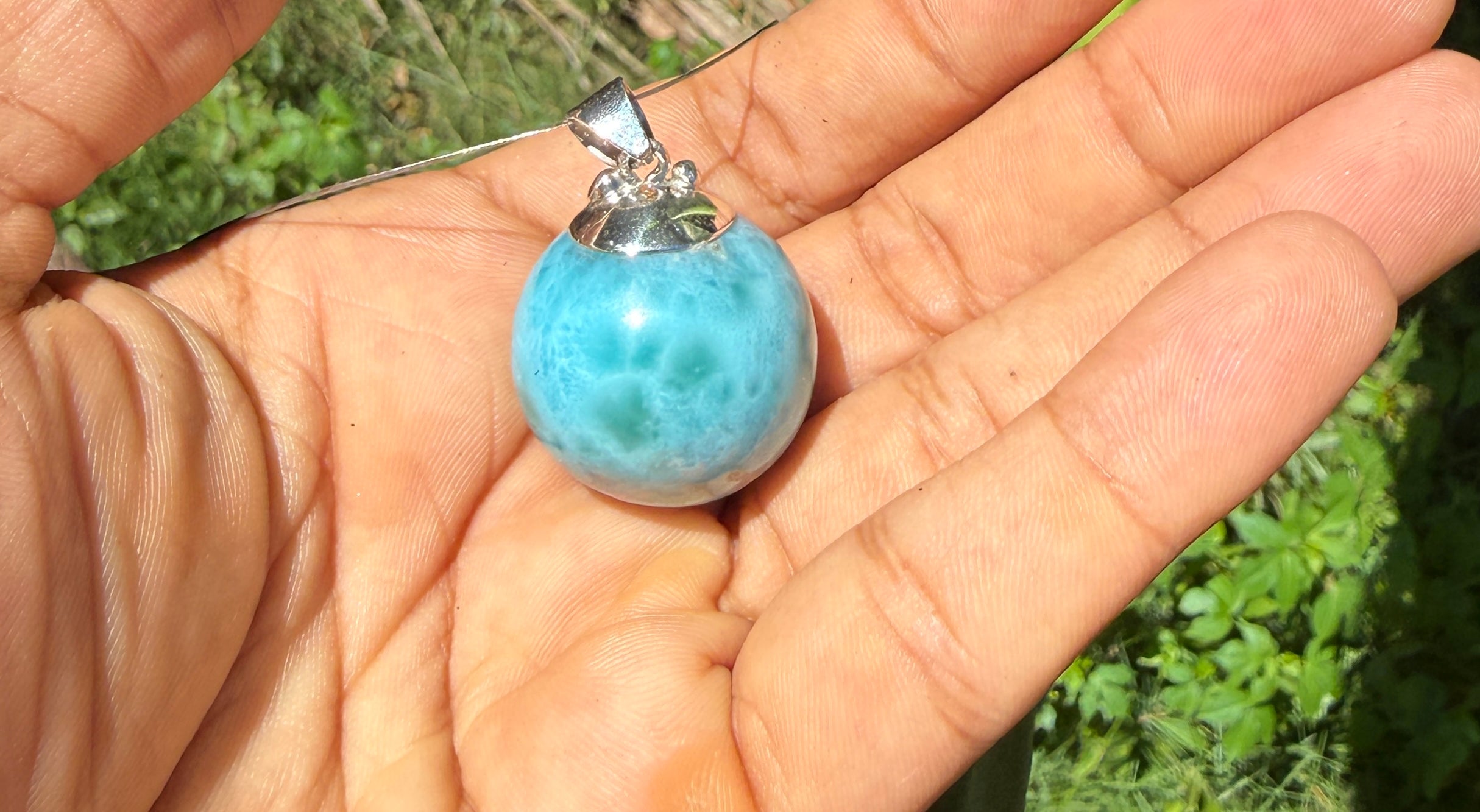 A light blue marbled larimar stone pendant with a silver bail, held in a person's open palm outdoors with green foliage in the background.