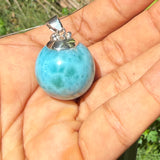 A light blue marbled larimar stone pendant with a silver bail, held in a person's open palm outdoors with green foliage in the background.