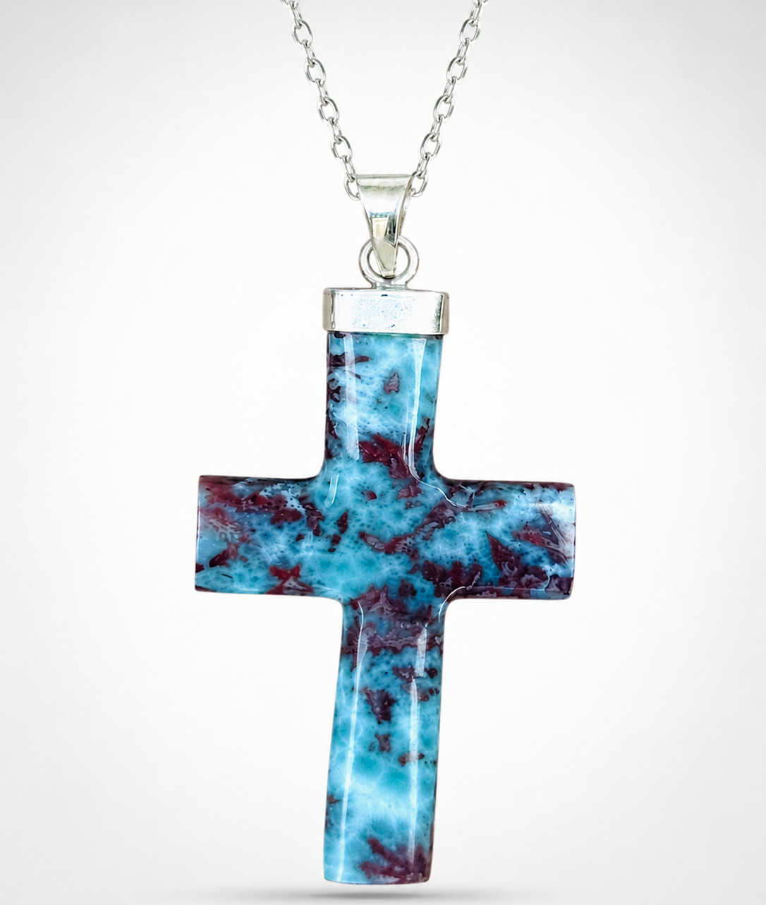 A silver Larimar cross pendant necklace featuring a polished turquoise and dark red marbled stone inlay, suspended from a silver chain against a light gray background.