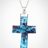 A silver Larimar cross pendant necklace featuring a polished turquoise and dark red marbled stone inlay, suspended from a silver chain against a light gray background.