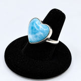 A heart-shaped Larimar ring with a silver bezel setting, displayed on a black velvet ring stand against a white background.