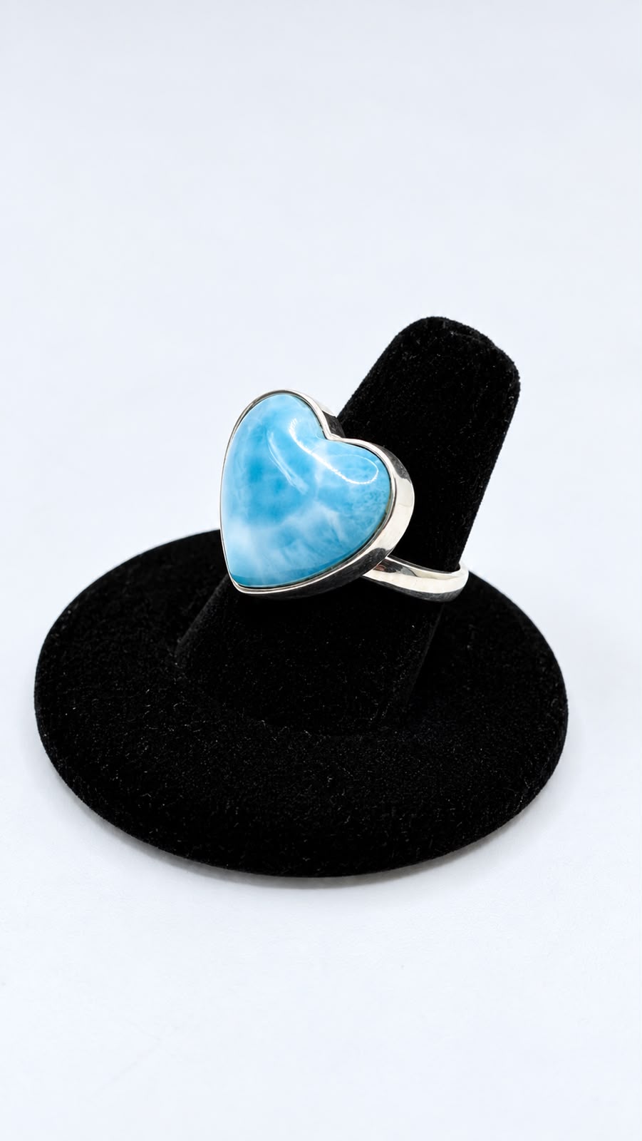 A heart-shaped Larimar ring with a silver bezel setting, displayed on a black velvet ring stand against a white background.
