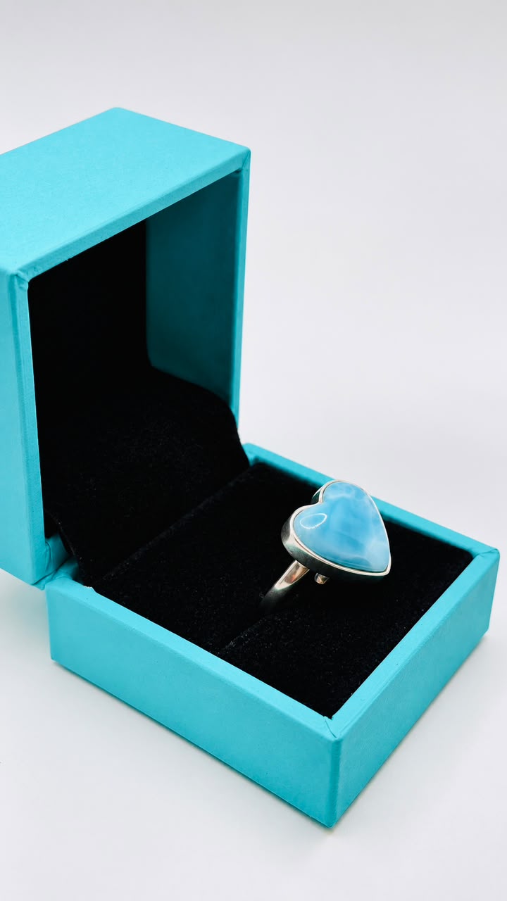 A silver ring with a heart-shaped light blue stone displayed inside an open turquoise jewelry box on a light gray background.