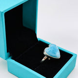 A silver ring with a heart-shaped light blue stone displayed inside an open turquoise jewelry box on a light gray background.