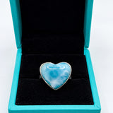 A heart-shaped ring with a light blue and white marbled stone in a silver setting, displayed inside an open turquoise jewelry box with a black velvet interior on a white background.