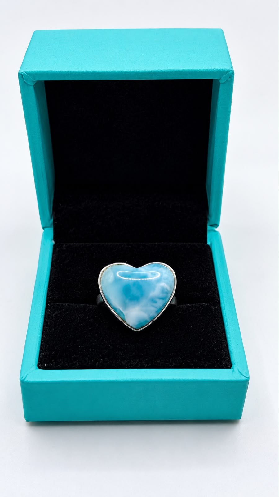 A heart-shaped ring with a light blue and white marbled stone in a silver setting, displayed inside an open turquoise jewelry box with a black velvet interior on a white background.