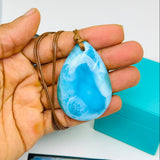 A hand holds a Larimar teardrop-shaped light blue marbled stone pendant on a brown cord necklace, with a turquoise box and a blue 'Mauna Vibes Certificate of Authenticity' card below on a white surface.