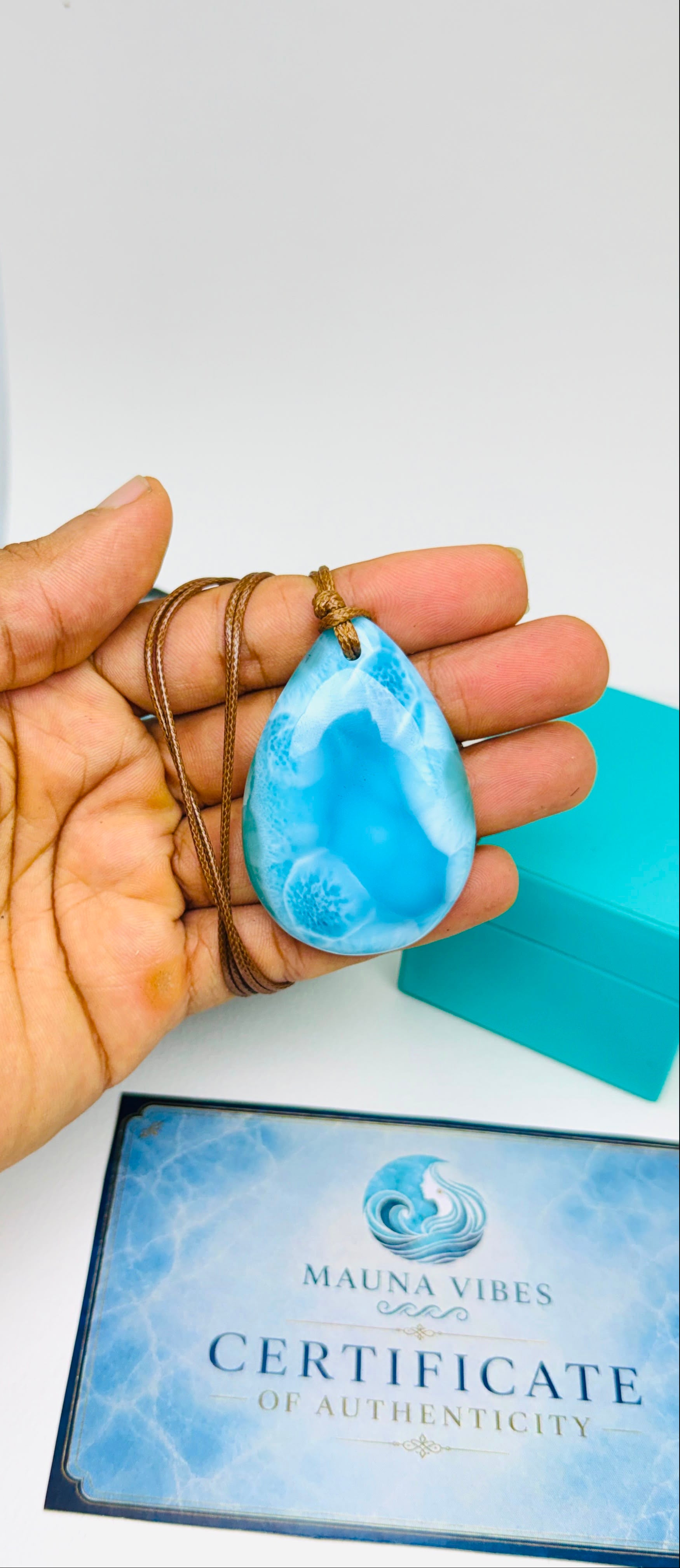 A hand holds a Larimar teardrop-shaped light blue marbled stone pendant on a brown cord necklace, with a turquoise box and a blue 'Mauna Vibes Certificate of Authenticity' card below on a white surface.