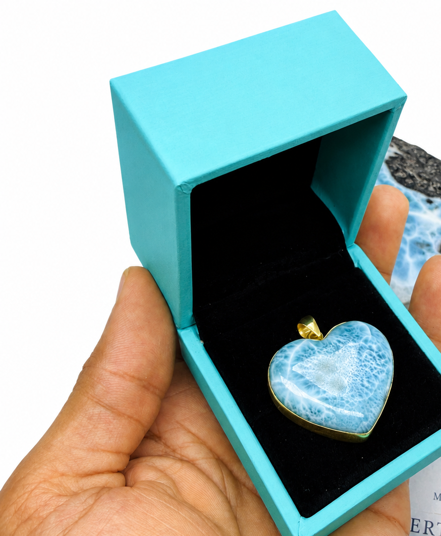 A heart-shaped blue  Larimar and white marbled pendant with a gold bail, displayed inside an open turquoise jewelry box held in a hand. A raw stone and a 'Mauna Vibes Certificate of Authenticity' card are visible in the background on a white surface.