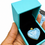 A heart-shaped blue  Larimar and white marbled pendant with a gold bail, displayed inside an open turquoise jewelry box held in a hand. A raw stone and a 'Mauna Vibes Certificate of Authenticity' card are visible in the background on a white surface.