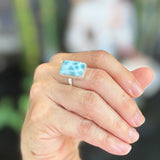 A close-up of a hand wearing a silver ring with a rectangular light blue larimar stone