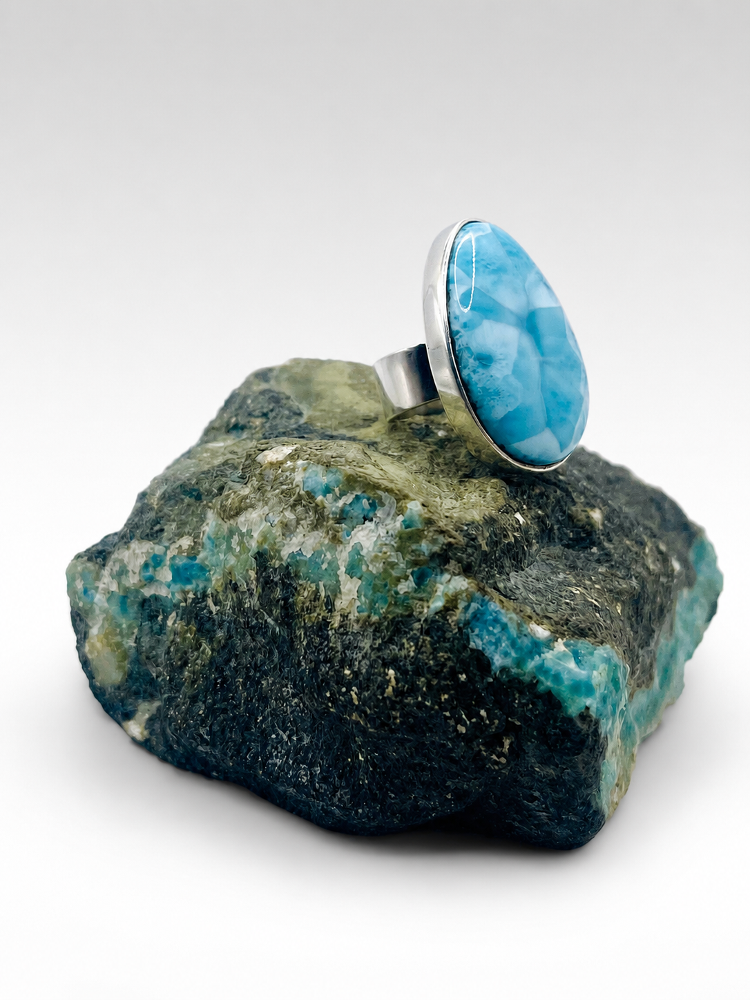 A sterling silver ring featuring a large, oval-shaped cabochon Larimar stone, displayed on a raw green and blue mineral specimen against a clean white background.