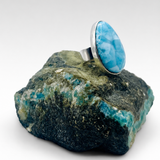 A sterling silver ring featuring a large, oval-shaped cabochon Larimar stone, displayed on a raw green and blue mineral specimen against a clean white background.