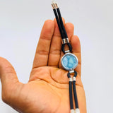 A hand holding a black cord bracelet with a round light blue stone in a silver bezel setting, accented with silver metal beads and a silver lobster clasp.