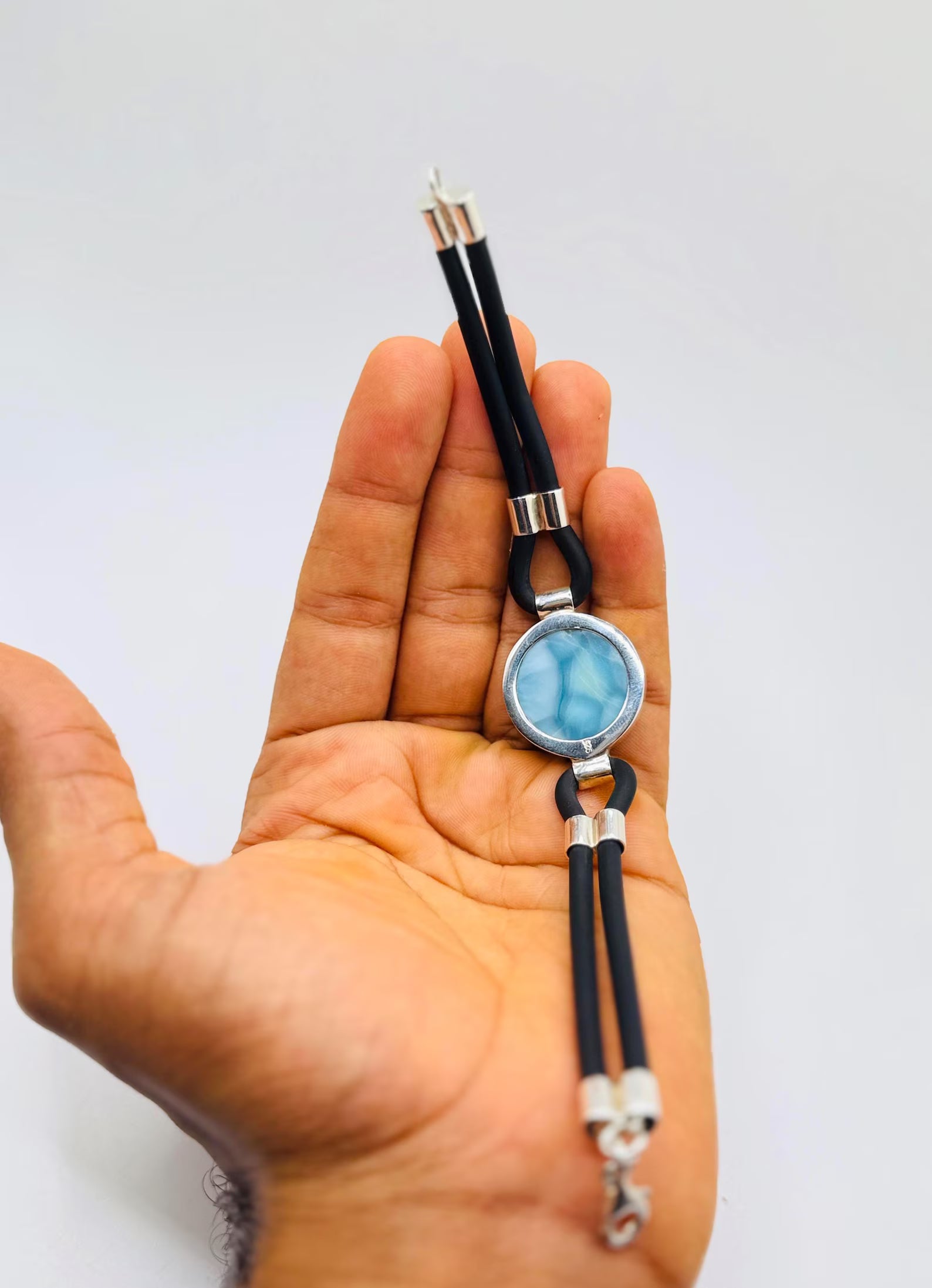A hand holding a black cord bracelet with a round light blue stone in a silver bezel setting, accented with silver metal beads and a silver lobster clasp.