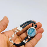 A hand holding a black leather bracelet with a round silver bezel containing a light blue marbled stone, alongside silver chain links and silver spacer beads.