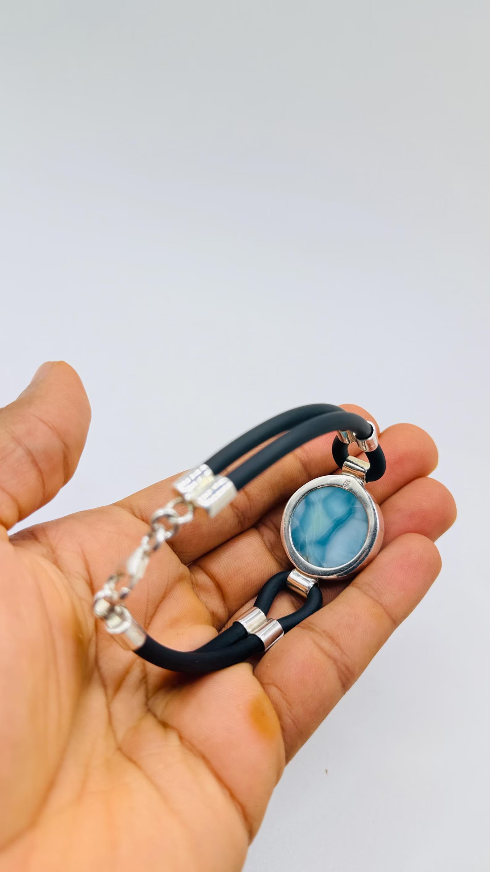 A hand holding a black leather bracelet with a round silver bezel containing a light blue marbled stone, alongside silver chain links and silver spacer beads.