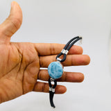 A hand holding a black larimar leather bracelet with a round light blue marbled stone charm and silver-toned metal accents, set against a plain light gray background.