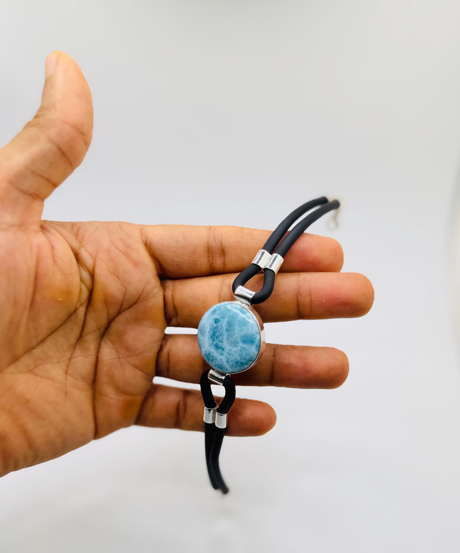 A hand holding a black larimar leather bracelet with a round light blue marbled stone charm and silver-toned metal accents, set against a plain light gray background.
