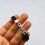 A black double-strand leather bracelet with a silver lobster clasp and a small silver charm, held in a person's hand against a plain light gray background.