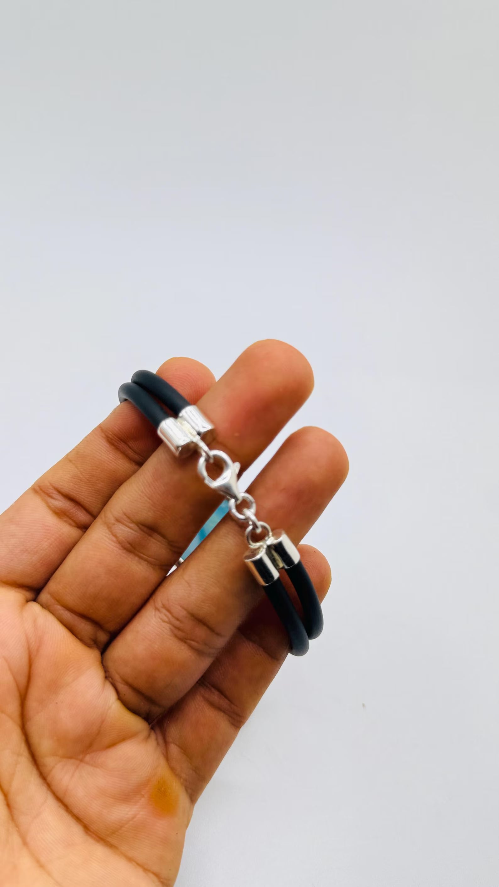 A black double-strand leather bracelet with a silver lobster clasp and a small silver charm, held in a person's hand against a plain light gray background.