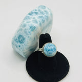 A silver ring with a round light blue and white marbled stone, displayed on a black stand next to a matching light blue and white marbled decorative object on a white background.