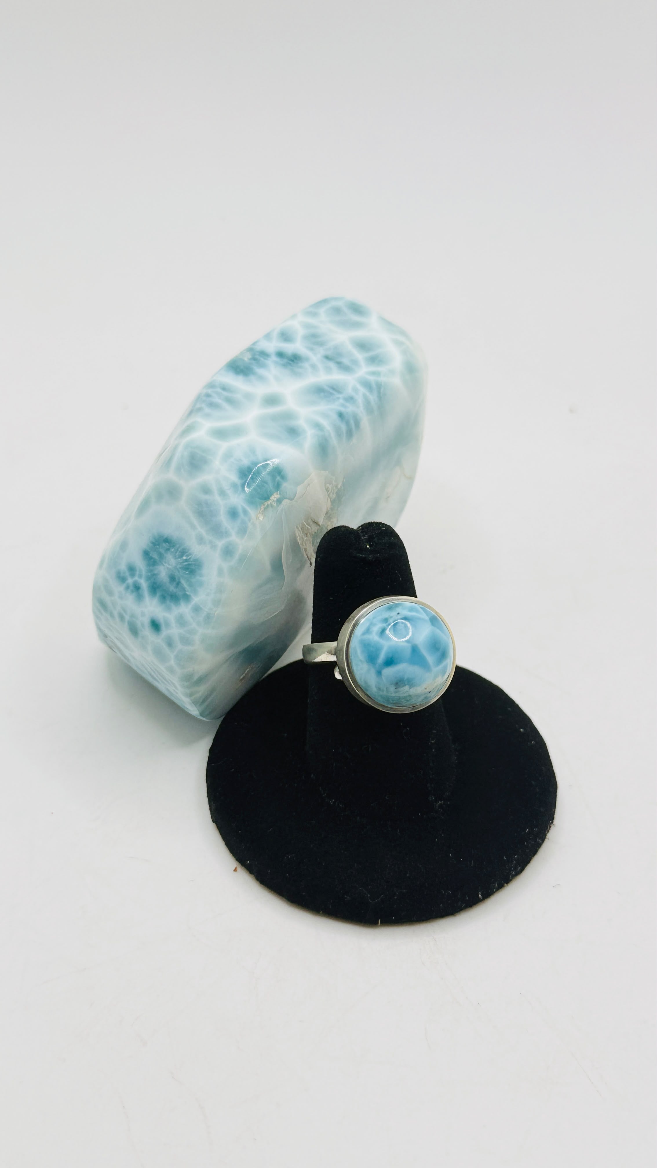 A silver ring with a round light blue and white marbled stone, displayed on a black stand next to a matching light blue and white marbled decorative object on a white background.