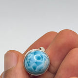 A silver ring with a round, domed blue larimar stone, held in a person's fingers against a neutral gray background.