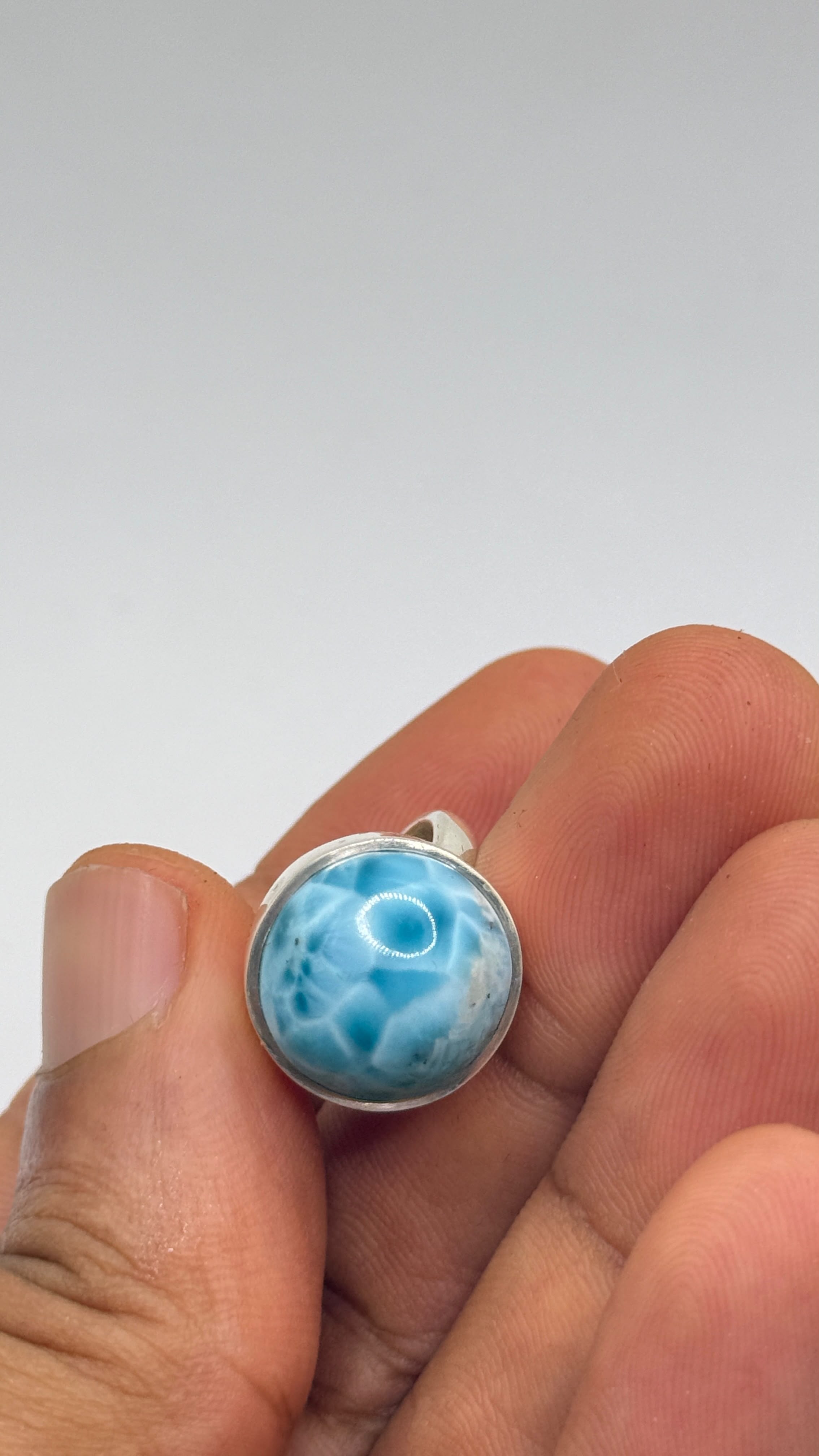 A silver ring with a round, domed blue larimar stone, held in a person's fingers against a neutral gray background.